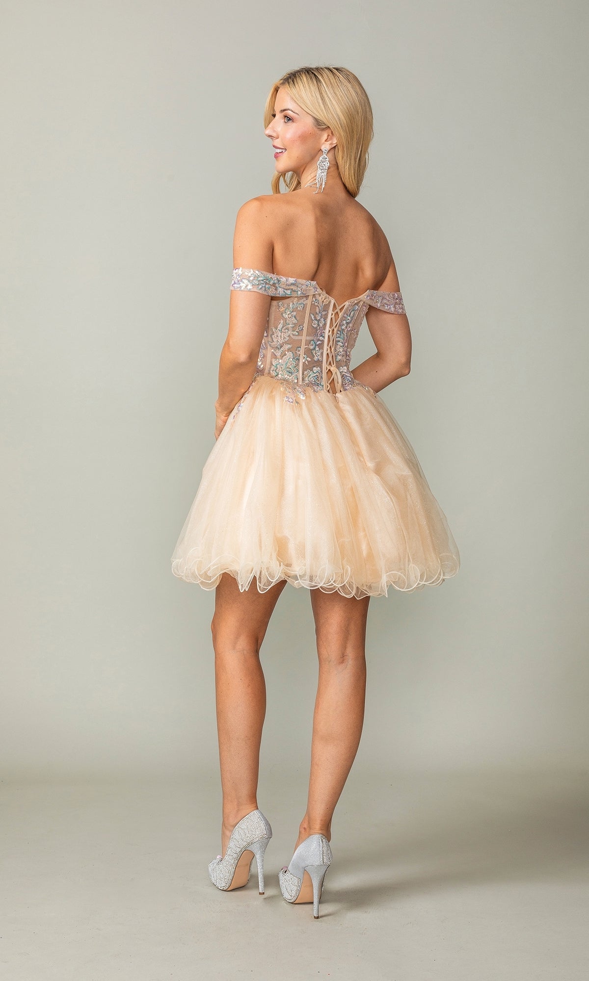 A woman faces away, wearing the Short Corset Homecoming Dress: Dancing Queen 3383 featuring a sparkly sheer corset bodice and full tulle skirt, paired with high heels and dangling earrings.